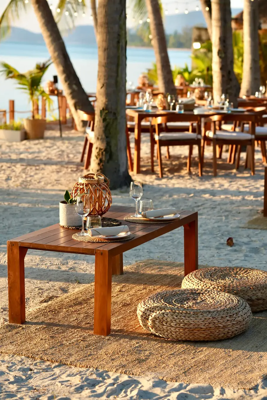 A table set for dinner on the sand beneath palm trees, lit by the low evening sun.